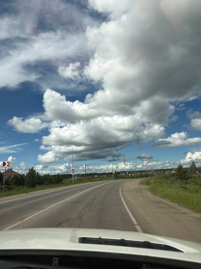The image depicts a curved asphalt road with white lane markings, viewed from the perspective of a vehicle moving forward. The road passes through a rural or suburban area with low-lying buildings and dense green trees lining the sides. Several traffic signs indicating curves are visible along the roadside. The sky is filled with a dramatic display of white and grey clouds against a bright blue backdrop. The scene suggests a daytime drive in a location like Severny, Russia, characterized by open roads and a dynamic sky.