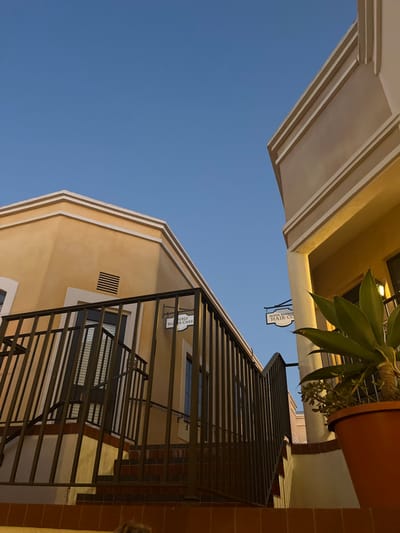 The image displays an exterior architectural scene within a commercial complex located at 121-123 East Anapamu Street, Santa Barbara, California. Two buildings constructed in a Spanish Colonial Revival style are visible, featuring light beige stucco facades with white trim and cornices, set against a clear, light blue sky.
In the foreground, a dark metal railing ascends alongside a set of steps constructed from reddish-brown tiles or bricks. On the left building, a white, rectangular hanging sign reads "ANGELS FOSTER CARE." On the right building, a similarly styled white sign, partially obscured by a large potted agave plant in a terracotta-colored pot, reads "SANTA BARBARA HAIR CO." The right building exhibits a warm, artificial illumination beneath its overhang. No individuals are present in the scene.