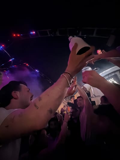 The image depicts a group of people in what appears to be a dimly lit gathering, potentially a party or celebration. The main focus is on an individual holding up a white cup or container, presumably offering it to others. Several hands are reaching towards the cup, suggesting a communal sharing of its contents. The environment seems to be indoors, possibly a bar or club, with colorful lights adding to the festive atmosphere. In the background, there's a ladder-like structure with lights on it, which might be part of the venue's decoration. It is impossible to determine from this image if the location is Olgiate Olona, Italy.