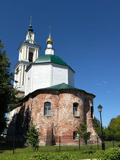 This is a low-angle, eye-level, full shot of the Church of the Resurrection of Christ in Vereya, Russia, on a clear, sunny day. The church is a historic brick building with white plaster elements. Its main body is made of weathered red brick, while the upper portions of the bell tower and the main dome are white with green roofs. The bell tower has a yellow and green spire with a cross. Another smaller dome with a gold cross is visible to the right of the bell tower. The church has arched windows, some with white trim. A black metal fence encloses the church grounds, and a black lamppost stands to the right of the building. Trees with green foliage are visible behind and to the left of the church. The sky is a deep, clear blue.