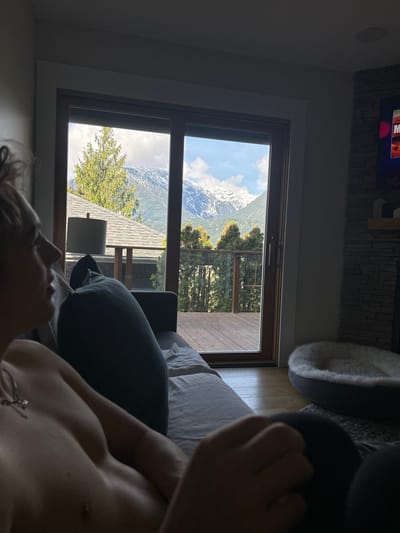 A first-person perspective shot shows a person lying on a couch in an interior setting. The primary view is through a large window showcasing a landscape with snow-capped mountains. Trees are visible in the middle ground and a rooftop of a nearby building is partially seen below. The location is likely Squamish, Canada given the mountain range. The person on the couch is partially visible, with an arm and a part of their torso. The lighting suggests daytime with sunlight entering the room.