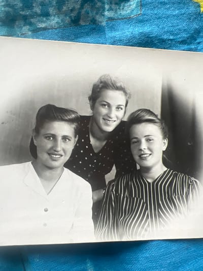This is a black and white photograph featuring three young women, likely friends, posing together. The woman in the foreground, wearing a white collared shirt, looks directly at the camera with a pleasant expression and a bright smile. Behind her, two other women are visible. The woman to her left has shoulder-length hair and is also smiling, looking slightly off-camera. She is wearing a dark polka-dot dress. The woman furthest back is wearing a striped top and is leaning her head on the shoulder of the woman in the polka-dot dress. She has blonde hair and is also smiling towards the camera. The women appear to be indoors, with a dark background that suggests a simple interior setting. The lighting in the photograph is soft, highlighting their faces. The overall mood of the image is cheerful and friendly. There is no visible text in the photograph. The context provided places the scene in Дружба Снт, Russia.