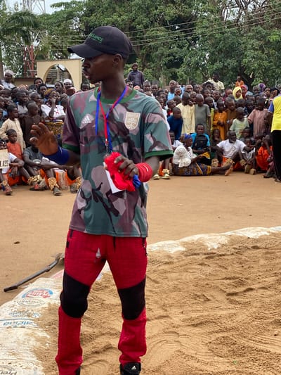 The image shows a man standing in a sandy area, possibly a wrestling arena. He wears a camouflage shirt with "AUSTIN" visible on the left breast and the Adidas logo on the right. He also wears red quilted pants with black accents and a black baseball cap with the word "RENAISSANCE" on it. He has a lanyard around his neck with blue and red stripes and is holding a red cloth in his left hand. His right arm has a blue wristband.
Behind the man, a large crowd of people is gathered. Some are seated, while others are standing. The crowd includes both men and women, and they are wearing various types of clothing. A white building is visible in the background behind the crowd.
To the right of the man is a large pile of sand and a partially visible white bag. There is a shovel leaning against the bag.
The image appears to have been taken in Kafanchan A, Nigeria, based on the clothing worn by people in the crowd. The scene suggests a cultural event, likely a wrestling match.