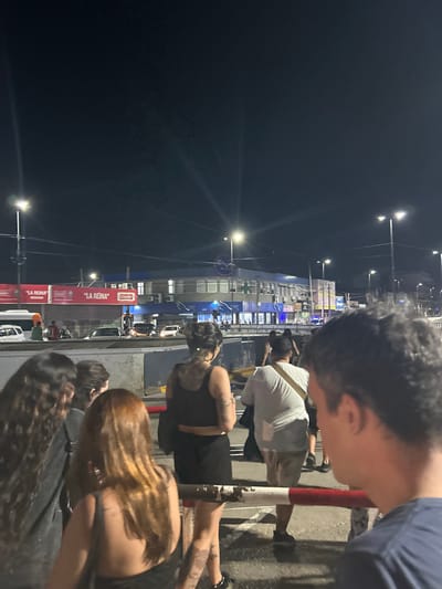 A group of people stands near a toll booth at night. Cars line up behind them. Streetlights illuminate the area. The sky is dark. A sign reads "LA REINA." The people appear to be diverse in age and style. A man with dark hair is visible in the foreground, looking down. The overall scene depicts a casual, public setting at a transportation hub.
