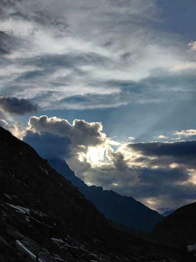The image depicts a dramatic, cloudy sky over a mountainous landscape in Damphug, India. Sunlight pierces through gaps in the thick, grey clouds, casting bright beams onto the scene. The foreground is dominated by the dark silhouette of a steep, rocky mountain slope. In the background, several other mountain peaks are visible, fading into the distance. A few small birds are present in the sky. The overall mood is one of natural grandeur and a dynamic interplay of light and shadow.