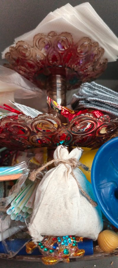 This is a close-up, indoor photograph of a collection of assorted items. In the foreground, a small, white drawstring bag made of textured fabric is tied with rough twine. Below the bag, a decorative metallic object, possibly a trinket or charm, is visible, adorned with colorful blue and red rhinestones and gold accents. To the left of the bag, a bundle of plastic drinking straws, striped in various pastel colors, is partially obscured by a clear plastic wrapper. To the right of the bag, a bright blue plastic funnel-shaped object is prominent. Behind these items, a red, ornate glass dish or bowl is visible, containing wrapped candies. Further back, a stack of gray fabric or material and a portion of white paper or napkins are present. The overall impression is of a somewhat cluttered, still-life arrangement. The image does not contain any discernible elements that would allow for the determination of its location in Tikhvin, Russia, or any other specific urban environment.