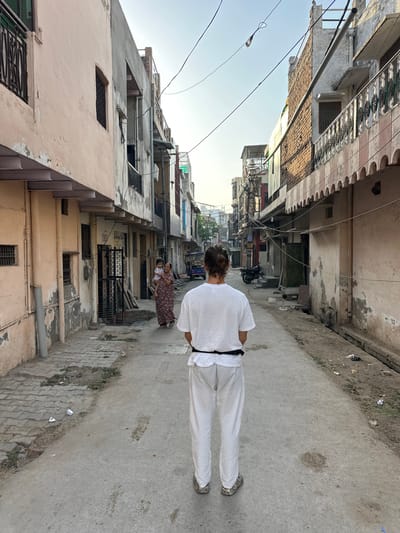 A street in Mathura, India, is depicted in a full shot. The street has light gray concrete on the right side and gray bricks on the left. Light gray concrete buildings line the street, with an older pink building on the left. Overhead, electrical wires traverse the street. A person dressed in a white outfit with a black belt walks away from the camera. In the background, an adult holds a child.