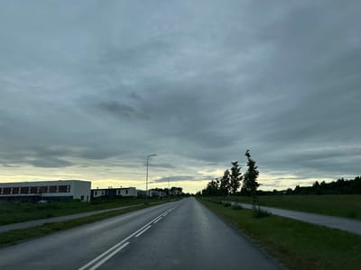 The image depicts a straight, asphalt road in Tiskre, Estonia, likely during twilight or overcast conditions given the dark clouds and subdued lighting. On the left side of the road, there are modern, multi-story residential buildings with white and dark facades, interspersed with grass and construction fencing. A sidewalk runs parallel to the buildings.

The road itself is divided by two white lines in the center. Several streetlights are visible along the left side of the road, stretching into the distance.

On the right side of the road, a paved pathway or another road runs parallel, bordered by a grassy field. A line of young trees, supported by stakes, is planted along the edge of the pathway. Further to the right, a dense line of dark green trees forms the background, with a patch of lighter sky visible above them. The overall atmosphere is calm and quiet, suggesting an early morning or evening scene in a suburban or developing area. There are no people or vehicles visible in the image.
