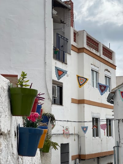 The image depicts a narrow urban street in Buñol, Spain, viewed from a slightly upward angle under an overcast sky. Two multi-story buildings, predominantly white with textured plaster facades, flank the scene.
On the left, a section of a white building features a vertical arrangement of five wall-mounted flower pots. From top to bottom, these are a green pot with a small green plant, a red pot with a purple-leafed plant, a blue pot containing a pink flowering plant, an orange pot with a succulent, and a partially visible green pot with a trailing plant. A white conduit runs vertically along this wall.
The larger building on the right is also white, distinguished by horizontal orange-brown bands at the first and second-story levels. The first story displays two windows with white frames; an external air conditioning unit is mounted beneath the left window. A small balcony with a dark metal railing is visible, with a green fabric item draped over it. The second story features a decorative parapet with a series of rectangular openings containing reddish-brown lattice work. Towards the top-left, a brick chimney with a television antenna is present against the sky. A dark, possibly grated, window or door is visible at the lower right.
Two lines of decorative, triangular crocheted banners are strung horizontally across the street between the buildings. The upper line includes triangles in light blue/orange/gray, yellow/black/light blue/gray, and maroon/blue/gray patterns. A lower line displays smaller, diverse yarn decorations, including a circular, multicolored piece with tassels. Black electrical wires are also strung across the street, some attached to the buildings. A gray downspout is visible on the far right building. No people are present in the scene.