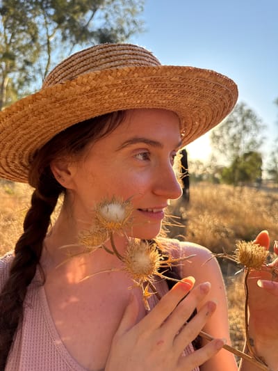 A young woman with auburn hair styled in a braid, wearing a straw hat and a pink top, is pictured in profile, looking to the right. She is holding two dried thistle flowers in her hands. The background is a sun-drenched field with out-of-focus trees and foliage, bathed in the warm light of what appears to be late afternoon. The scene suggests an outdoor setting, possibly in the rural outskirts of Katzrin, Israel, where such dry vegetation and open fields are common.