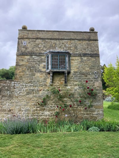 The image depicts a weathered stone wall, likely part of an old building or structure. The wall is constructed from irregularly shaped stones, with visible mortar lines. The image is taken in Sevenoaks, United Kingdom. A square window is set into the wall, with a greenish frame and multiple panes. A climbing rose bush with red flowers is growing on the right side of the wall, partially obscuring the stone surface. To the right of the wall, there is a strip of green grass, suggesting a garden or park setting. Above the window, there are metal numbers, the number 18. The top of the wall features a decorative ledge and two spherical objects at the corners. The sky is cloudy, with a soft light filtering through. The building is perpendicular to the horizon.