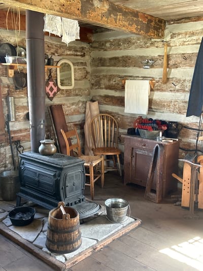 This image depicts the interior of a rustic log cabin, likely a historical exhibit or a preserved living space. The walls are constructed of rough-hewn logs with chinking between them. A dark, ornate cast-iron wood stove stands prominently in the foreground, with a pot resting on its top and a bucket of kindling to its side. To the left of the stove, a metal bucket is visible.

The room is sparsely furnished with a mix of wooden furniture. A simple wooden chair with a woven seat is positioned near the stove, and another wooden chair, this one with a spindle back, is further to the right. A small, weathered red cabinet with a drawer and a door stands against the back wall, topped with a bowl of colorful yarn, a pitcher, and a candleholder. Above the cabinet, a white cloth is hung from a wooden rack.

Various objects are scattered throughout the cabin, suggesting a lived-in or functional space. Shelves on the log wall hold metalware, a small mirror, and a decorative quilt square. A broom is leaning against the wall, and other tools and containers, including wooden buckets and metal pails, are visible. The ceiling beams are rough and unfinished, and some laundry is hung to dry from one of them.

The floor is made of wide wooden planks. Sunlight streams in from an unseen source, casting bright patches and shadows across the room, highlighting the textures of the wood and the dust motes in the air.

This scene is consistent with the interior of the Stormont, Dundas and Glengarry Historical Society's historic building exhibit, possibly the Van Allan House, located in the city of South Dundas, Ontario, Canada. The exhibit aims to showcase pioneer life in the region.