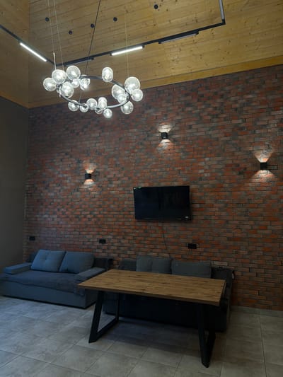 The image shows an interior space with a mix of textures and materials. The ceiling is divided between wooden planks and a brick wall. A modern chandelier hangs from the wooden ceiling. The brick wall features a flat-screen TV and four square sconce lights. A grey sectional couch and a table with a wooden top and black metal legs are positioned near the brick wall. The floor is covered in grey tiles. The location is possibly Kamennoye, Russia, based on the prompt. No people are visible.