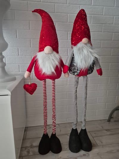 The image shows two decorative gnomes standing in front of a white brick wall. The gnome on the left is wearing a red sequined hat, a red tunic, and striped red and white leggings. It is holding a red sequined heart in its left hand. The gnome on the right is wearing a red sequined hat, a gray vest with silver sequins, and gray ribbed leggings. Both gnomes have white beards and pink noses. They are standing on a wooden floor. The background consists of a white brick wall and a white cabinet on the left side of the image. There is no indication that this scene is located in Barbing, Germany.