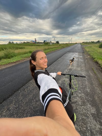 This is a first-person perspective, vertical media file taken outdoors. The primary subject is a woman riding a bicycle. She is wearing a black and white bomber-style jacket with striped cuffs. Her hair is dark brown and pulled back, and she is smiling at the camera.
The setting appears to be a rural road, possibly in Pivkino, Russia, given the location context. The road is paved with asphalt, and there is a strip of freshly laid asphalt alongside it, suggesting recent road work. Green grass and vegetation border the road, and in the distance, there are some trees and buildings. A dog is visible in the middle of the road ahead of the cyclist.
The sky is dramatic, with heavy clouds that suggest either an approaching storm or recent rain. The light is soft and diffused, indicating it is likely early morning or late evening.
The overall mood of the image is cheerful and active, with the woman enjoying a bike ride. There is no visible text in the media file.