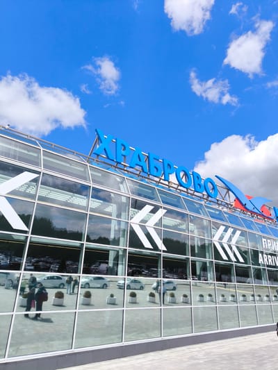 The image captures a modern glass-fronted airport terminal building under a bright blue sky with scattered white clouds, indicating clear daytime weather. The setting is Khrabrovo, Russia, as prominently displayed by large blue Cyrillic letters spelling "ХРАБРОВО" (Khrabrovo) on the roof of the building. To the right of this sign, a blue and red logo, possibly stylized wings or an airplane, is visible.

The building's glass facade reflects the surroundings, including a paved forecourt, cars, and several people. White arrow-like graphics are applied to the glass panes, possibly indicating direction or decorative elements. Further down on the glass, partially visible text reads "ПРИЛЕТ ARRIVAL," suggesting an arrivals area.

Reflected in the lower section of the glass, several individuals are seen walking on the pavement outside. Some appear to be carrying bags, consistent with airport activity. A line of white potted plants with green shrubs is also reflected. Behind the reflections of the people and plants, numerous cars are visible, either parked or in motion, implying a busy drop-off or pick-up area. A security camera can be spotted on the roofline. The overall scene depicts a functional, active airport environment.