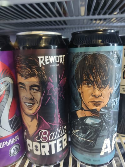 Two cans of Rewort brewery beer are stacked on top of each other in what appears to be a refrigerated display rack. The top can has an image of a person and a motorcycle, with a light blue background. The bottom can has an image of a person and the words "Baltic PORTER" in white text with a purple outline, on a black background. The name "REWORT BREWERY" is visible on both cans. The cans are in a wire rack, likely in a store or cooler. The location is Pskov, Russia, based on the Cyrillic text visible on the can underneath the Rewort Baltic Porter.