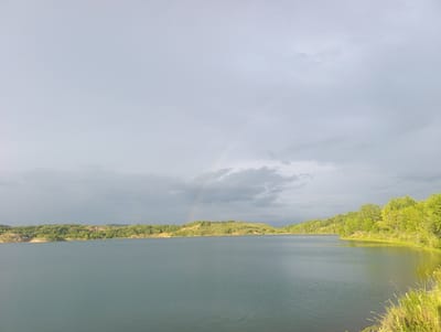 The image depicts a tranquil lake scene under a cloudy sky, hinting at recent rain or the possibility of more to come. A faint rainbow arcs across the sky, suggesting a break in the clouds. The water is a deep, muted blue, reflecting the overcast sky. Lush green trees and vegetation line the shores of the lake, creating a natural and somewhat wild landscape. The terrain appears hilly and uneven, with some exposed earthy banks visible on the left side of the image, contrasting with the dense greenery elsewhere. The time of day is indeterminate due to the cloud cover, but the lighting suggests it could be late afternoon or early evening. The overall mood is peaceful and serene, emphasizing the natural beauty of the location. There are no visible people, objects, or text in the image. The scene is characteristic of a natural landscape in Yaroslavsky, Russia, with its characteristic rolling hills and bodies of water.