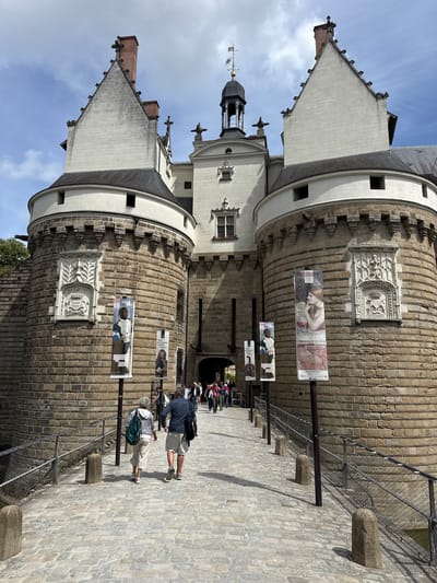 This image shows the entrance to the Château des Ducs de Bretagne in Nantes, France, on a bright, cloudy day. A cobblestone bridge leads to the castle's fortified entrance, flanked by two large, round stone towers. The towers have ornate stone carvings above arched doorways, and banners advertising an exhibition are displayed on poles along the bridge.
Several people are walking towards the castle entrance. In the foreground, a couple is walking away from the camera, the woman with a green backpack and the man with a dark backpack. Further ahead, a group of people are seen entering the castle. The castle itself is built of light-colored stone and features steep, pointed roofs and a central clock tower. The overall scene depicts a historical landmark bustling with visitors.