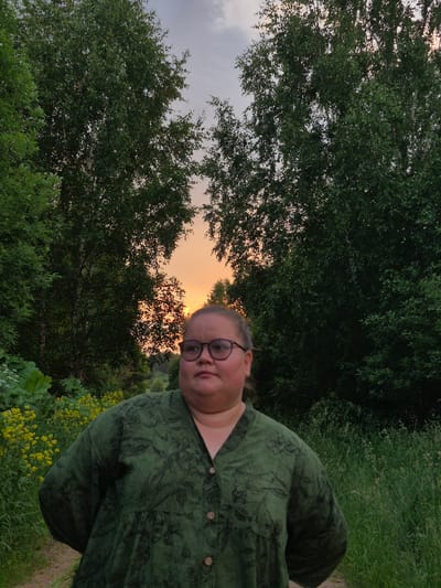 A woman with glasses and a green dress stands in a forest during sunset. The sky is orange and purple, and the trees are green and lush. The woman is looking to the side, and her hands are behind her back. She appears to be standing on a dirt path, with tall grass and yellow wildflowers on either side. The location is Doinaya, Russia.