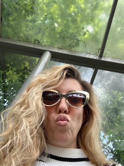 A woman with long blonde wavy hair and sunglasses is posing with puckered lips in what appears to be an enclosed bus shelter or waiting area. The structure has large, somewhat dirty windows revealing green foliage outside, possibly trees. A metal handrail is visible near her. She is wearing a striped shirt. The location is unknown.
