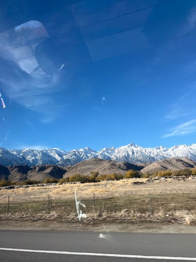 This media file captures a vast, scenic landscape, oriented as if taken from a vehicle looking across a road towards mountains under a bright sky. The upper half of the image is dominated by a clear, deep blue sky, featuring a few wispy white clouds and faint contrails.
A majestic range of rugged snow-capped mountains stretches across the middle of the frame. Their peaks and upper slopes are covered in bright white snow, contrasting with the darker, rocky lower sections. Along the base of these mountains, a horizontal band of trees or bushes displays yellowish-brown foliage, indicating either autumn or winter conditions.
Below the treeline, a dry, arid-looking grassland or scrubland extends, making up the immediate foreground and the terrain leading up to the mountains. On the far right, a paved road or shoulder is visible, with a clear white line demarcating its edge.
The left portion of the image, particularly the sky, shows prominent reflections, suggesting the photograph was taken through a window, likely a car's side window. These reflections include several horizontal parallel lines, resembling power lines or window frames, and indistinct, swirling patterns, possibly from smudges or internal reflections. A notable bright, curved reflection is visible in the upper-left sky area.
The time of day is bright and sunny, under clear weather conditions. The presence of snow on the mountains and dry vegetation suggests late fall, winter, or early spring. No individuals, animals, or visible text are present. The scene evokes a sense of natural grandeur and open spaces, consistent with the context of Lone Pine, United States.
