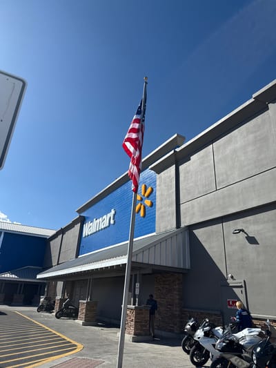 In Fernley, United States of America, an American flag waves in front of a Walmart store. The store has a distinctive blue facade with the Walmart logo prominently displayed. The exterior of the building is primarily gray with architectural detailing. Several motorcycles are parked in front of the store, suggesting a point of sale. Two individuals are visible near the entrance, one standing and the other seated. The sky is clear and blue, indicating fair weather conditions. A parking lot with yellow lane markings is in the foreground, and a portion of a white sign is visible on the left side of the frame.