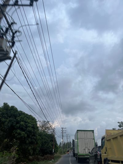 The image depicts a roadside scene in Payarangkuluh, Indonesia, under a predominantly overcast, grey sky. In the upper left, a utility pole with multiple transformers and a complex array of black electrical wires extends diagonally and horizontally across the sky, converging towards the distant horizon. The asphalt road, which appears to be a two-lane or shoulder-lined route, recedes into the background. On the left side of the road, dense green trees and foliage line the path, with some ground debris visible at their base. On the right side of the road, a line of traffic is observed moving away from the viewer. This traffic includes the rear of a yellow heavy-duty dump truck in the foreground, followed by a black SUV, and a large green and white box truck further down the road. A lone individual is operating a motorcycle on the left side of the road, also traveling away from the viewer, positioned between the trees and the moving trucks. Additional utility poles with multiple cross-arms are visible along the right side of the road in the mid-ground distance. The overall lighting is subdued due to the extensive cloud cover.