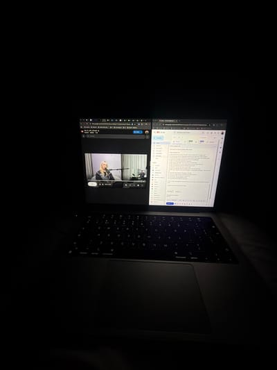 In a dimly lit room, a laptop is open, displaying two browser windows side-by-side. The left window shows a video player with a woman speaking into a microphone, possibly from a podcast or interview. The right window displays a Gmail inbox and an email composition screen. The keyboard of the laptop is visible in the foreground, suggesting someone is actively using the device. The overall environment appears to be indoors, likely a home or office, with no discernible features indicating a specific location in Ridderkerk, Netherlands.