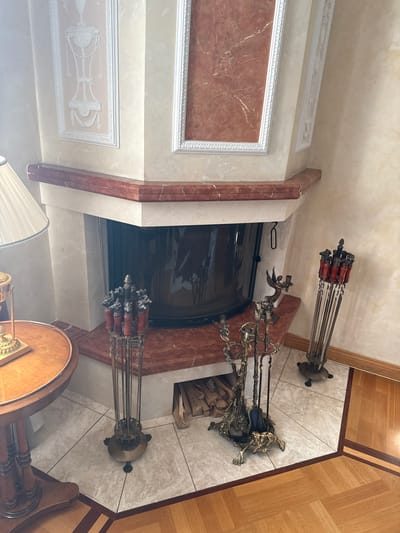 The image shows an interior scene with a fireplace. The fireplace has a marble mantelpiece with a decorative red marble inlay. To the left of the fireplace, a table with a wooden top and decorative metal base is visible, with a lamp on top. In front of the fireplace, there are several fire tools, including a set of tongs, a shovel, and a poker, displayed in a decorative stand. Another set of fire tools is visible to the right, also in a stand. The floor is tiled with a marble-patterned tile. The walls are decorated with ornate plasterwork. To the right, a section of wooden flooring is visible. The scene appears to be located in a residential building in Yukki, Russia, likely a living room.