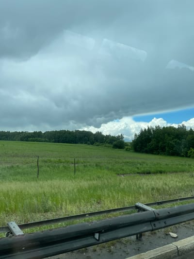 A roadside view presents a stark contrast in weather conditions. To the left, heavy, dark storm clouds dominate the sky, suggesting impending rain. To the right, there are fluffy white clouds against a blue sky. A lush green field stretches towards a tree line that separates the sky from the grassy area. A guardrail, composed of metal posts and rails, runs parallel to the road, indicating a raised or sloped embankment. The overall environment is rural or suburban, with a focus on the natural landscape juxtaposed against the manufactured safety barrier. The image does not contain any people. There is no definitive confirmation that this image is related to Кокино, Russia.