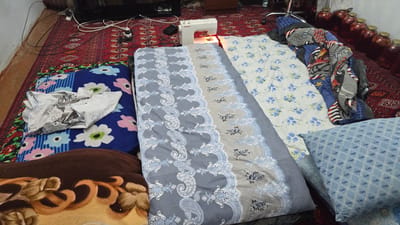 In a room in Kaka, Turkmenistan, a sewing machine sits on a patterned rug. Several rolled-up mattresses and pillows are arranged on the floor, covered with blankets and fabric. A sewing machine, identified by the brand name "JANOME" visible on its casing, is positioned on the rug near the mattresses. Various fabrics and materials, including a blue and white floral patterned fabric and a brown blanket with an ornate design, are laid out. A pile of rolled-up blankets and scarves, featuring various patterns like racing flags and stars, is also present. In the background, jars filled with what appear to be preserved fruits are lined up on a shelf. The overall scene suggests a domestic setting where sewing and domestic activities might be taking place. The lighting appears to be indoor lighting. There are no visible people in the image.