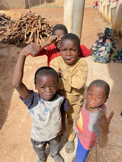 An overhead photograph captures a vibrant outdoor scene in Rahwol Chom, Nigeria, featuring a group of playful children and an adult under bright, direct sunlight. The scene is set on a surface that mixes packed dirt with lighter-colored concrete or tiled areas.
In the foreground and midground, four young boys are the primary subjects, looking up at the camera with cheerful expressions. The boy closest to the camera on the right wears a light t-shirt adorned with blue dinosaur patterns and dark pants; he smiles broadly with a raised fist. Beside him, another boy in a tan long-sleeved shirt playfully sticks out his tongue while smiling. To his right, a third boy wears a grey and reddish-pink t-shirt featuring a rainbow graphic and blue pants; he smiles and makes a peace sign with his right hand. Visible text on his shirt reads "GRIMUS S". A fourth boy, partially visible on the left, wears a red top. All the children have a light-colored residue, possibly dust or flour, on their faces.
In the background, to the upper left, an adult, likely female, is seen kneeling or squatting with her back to the camera. She is dressed in a brightly patterned traditional garment, possibly made from Ankara fabric, featuring green, blue, and white designs, and her hair is styled in long braids. To the lower left, a large pile of neatly stacked cut wood or branches, likely firewood, is visible.
Strong shadows suggest the photo was taken on a sunny day, likely around midday. In the far distance on the left, three small, indistinct figures can be seen standing near a building. The children's expressions convey happiness and youthful energy, indicating a moment of playful interaction with the photographer.