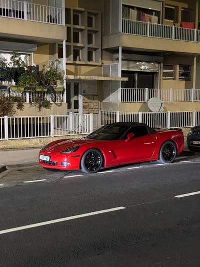 A red Chevrolet Corvette C6 convertible, with black multi-spoke wheels and a black soft top, is parked parallel to a curb on an asphalt street at night. The vehicle's front license plate displays "302387 NC." A portion of a dark-colored SUV is visible parked behind the Corvette on the right, with a partially visible license plate beginning with "449 02." The street surface features white dashed line markings.
In the background, a multi-story residential building with a light yellow-beige facade is illuminated by artificial light. The building has multiple balconies equipped with white metal railings. On the first-floor balcony to the left, various potted plants and a bicycle are visible. Below this, a ground-floor entrance features a series of mailboxes to the left of the main door. On the right side of the building, a white satellite dish is mounted on the facade below an upper-level balcony where laundry items are hanging to dry. No individuals or specific actions are discernible. This scene is located in Noumea, France.