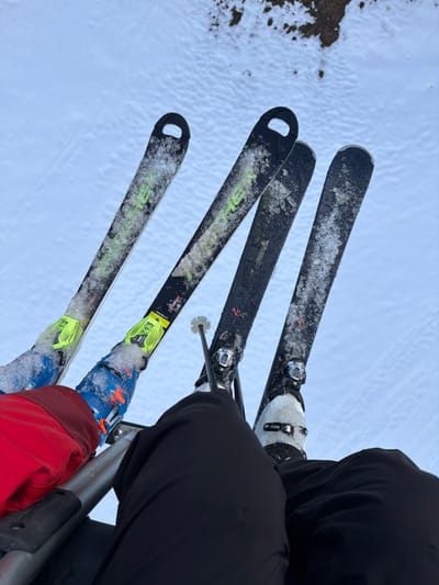 A high-angle view from a ski lift in Zweisimmen, Switzerland, shows the lower torsos and ski equipment of two individuals suspended above a snow-covered landscape. The person on the left wears a red jacket, blue Fischer ski boots with yellow bindings (marked "13"), and black skis with green accents and tips. The person on the right wears black ski pants, white ski boots, and black skis, one of which features the marking "N14". A black ski pole with a white basket is held by the right individual, positioned between their skis. All visible skis and boots are coated with accumulated snow and ice. A metallic safety bar from the ski lift is visible horizontally in the lower foreground. The ground below is predominantly covered in white snow, with a patch of darker, exposed earth visible in the upper right background.