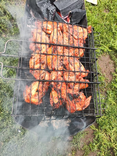 The image shows chicken pieces cooking on a barbecue grill over hot coals. The chicken is seasoned and appears to be well-cooked, with some charred edges. Smoke rises from the grill, obscuring some of the food. The grill is placed on a black metal barbecue stand, which is set against a background of green grass and dirt. There are also some pieces of charcoal visible in the barbecue. The scene suggests an outdoor gathering or meal, likely a summer barbecue given the green grass. The overall impression is one of a casual, rustic outdoor setting. The image appears to have been taken during daylight hours, with natural light illuminating the scene. There are no people or text visible in the image.