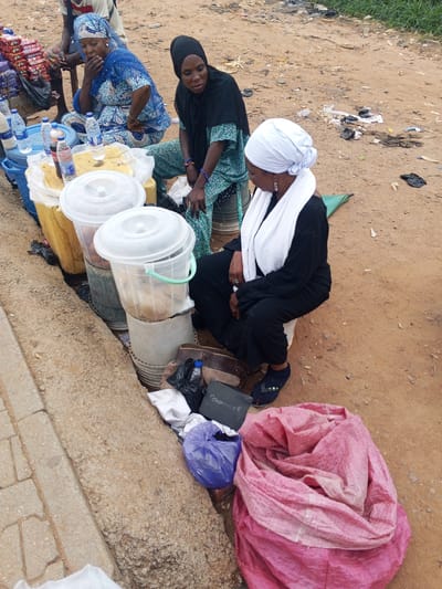 In Immigration Quarters, Nigeria, three women are seen in an outdoor market scene. One woman, seated on the ground, wears a blue and white patterned dress and a blue headscarf, with her hand resting on her mouth. Beside her, another woman in a green and blue patterned garment and a black headscarf is seated and looking down. A third woman, wearing a black outfit and a white headscarf, is also seated, facing away from the viewer, with her hands resting on her lap. In the foreground, several large, clear plastic buckets with white lids are stacked, containing a light-brown granular substance. Various items, including bottles of water and other beverages, are arranged on a yellow surface next to the buckets. A large, pink woven bag lies on the ground in the lower right corner of the image, and other bags and miscellaneous items are scattered around the scene, including a dark grey bag with visible text. The ground is composed of dirt and gravel, with some patches of dry grass and debris visible in the background. A concrete curb lines the left side of the frame.