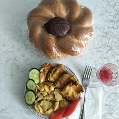 The image displays a still life composition on a marble-patterned tabletop, featuring a meal and a braided bread with a chocolate topping. The meal is served on a white plate with a brown rim and consists of fried potatoes, what appear to be battered zucchini slices, cucumber slices, and two wedge-shaped pieces of a red vegetable, likely tomato. A silver fork rests on a white paper napkin to the right of the plate. Beside the napkin is a small, ornate glass filled with a pink liquid, presumably a beverage. Above the plate, a large, braided loaf of bread, enclosed in clear plastic wrap, is visible. A single, dark brown, oval-shaped chocolate confectionery is placed centrally on top of the bread. The overall lighting suggests an indoor setting, possibly a home environment. There are no people present in the image. The scene is presented from a top-down perspective. The image is a photograph, and there is no information or visual cue within the image itself that specifies a location in Cupcini, Moldova.