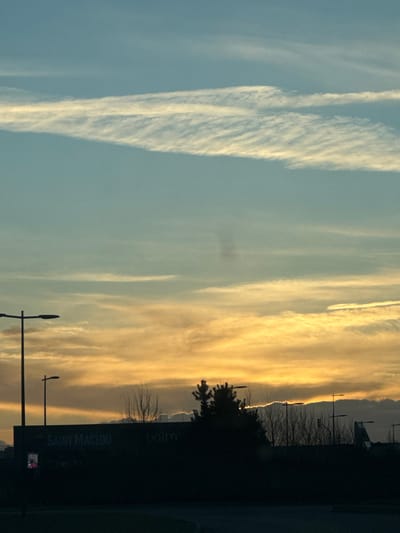 The image showcases a sky divided into blue and golden sections by linear clouds. On the left, streaks of white clouds stand out against the blue sky. The right part of the image shifts to golden hues due to the setting sun, with softer cloud formations. A silhouetted landscape runs along the bottom, including trees, buildings, and streetlights, and a sign which reads "SAINT-MACLOU". The photo is taken from a low perspective, looking upward.
