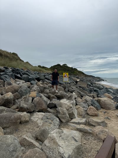 The image shows a man standing on a rocky shore in Courtown, Ireland. The man is wearing a dark t-shirt and shorts, and is looking down. He is standing amidst a large collection of grey and brown rocks that form a barrier along the coastline. In the background, there is a beach with gentle waves and a cloudy sky. A yellow warning sign with black text and symbols is visible to the right of the man, indicating a hazard. The sign is positioned on a metal pole and appears to be in Irish and English, warning of danger and advising to "Keep off the rocks." The overall scene depicts a rugged coastal environment on an overcast day.