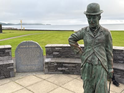 This media file features a bronze statue of Charlie Chaplin in Waterville, Ireland. The statue depicts Chaplin in his iconic Tramp persona, complete with a bowler hat, suit, and cane. He is positioned in the foreground, looking slightly to the left, with a stone monument to his left and a stone wall with a bench behind him.

The monument is inscribed with text honoring Charlie Chaplin, detailing his connection to the area and acknowledging the creators and funders of the statue. Visible text includes "CHARLIE CHAPLIN," "FEAR NA SCANNAN CARA AN CHOIREAIN A CHAITH BLIANTA SHONA INAR MEASC FOR THE MAN WHO MADE THE MOVIES SPEAK IN THE HEARTS OF MILLIONS CHARLIE SPENT MANY YEARS IN OUR MIDST AS A WELCOME AND HUMBLE GUEST AND FRIEND TO MANY. THIS IMAGE WAS CREATED BY SCULPTOR ALAN RYAN HALL IT WAS FUNDED BY THE GENEROSITY OF JOSEPHINE CHAPLIN AND BY THE EU LEADER PROGRAMME. I.R.D. WATERVILLE, OUR LOCAL COMMUNITY ORGANISATION INITIATED THIS PROJECT WHICH WAS UNVEILED IN JULY 1998."

The setting is outdoors, with a grassy lawn stretching towards a body of water, likely the Atlantic Ocean, and hills in the distance. The sky is overcast, suggesting a cloudy or misty day. The overall mood is one of remembrance and tribute. The statue is a prominent object in the scene, serving as a focal point for visitors. The presence of the monument and the statue together creates a memorial space dedicated to the beloved entertainer.