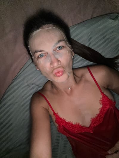 A woman with blue eyes and dark hair pulled back is lying on a bed. She is wearing a sheet mask and a red lace camisole. She is puckering her lips and looking towards the camera. The room is dimly lit. This scene is likely indoors, in a bedroom. No details about the city of Lapčići, Montenegro are present in the image.