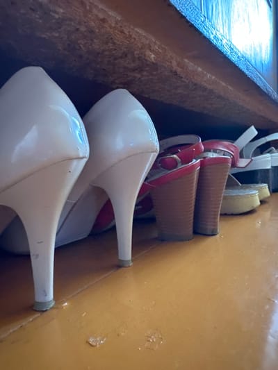The image displays a low-angle view of several pairs of women's high-heeled shoes stored on a wooden shelf. In the foreground, two light-colored patent leather pumps are visible, with their pointed toes facing the viewer. To their right, a pair of red and tan platform sandals with ankle straps is positioned. Further to the right, a pair of white and black sandals with a wedge heel and a light-colored sole is partially visible. The shoes are resting on a floor that appears to be made of polished wood, with some small, reflective wet spots and debris scattered across its surface. The shelf above is made of dark wood with visible grain. The overall impression is of shoes being stored indoors. There is no specific indication of the city of Urazovo, Russia within the image itself.