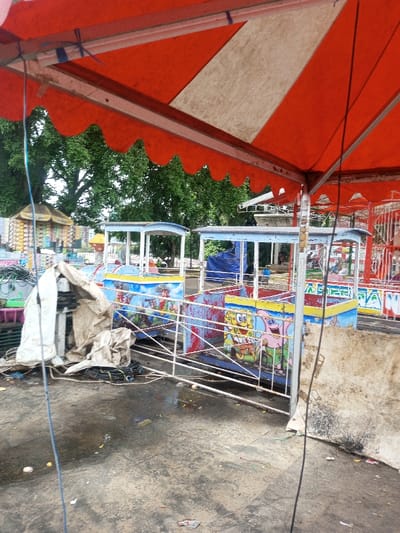 A rundown amusement park ride is the focal point, its small train carriages adorned with faded children's cartoon characters. The ride is fenced off, its appearance suggesting abandonment or disrepair. Above, a red and white striped awning offers partial cover. The ground is damp and littered, reinforcing the neglected atmosphere. The scene is overcast, with green trees visible in the background, hinting at a park setting beyond the ride.