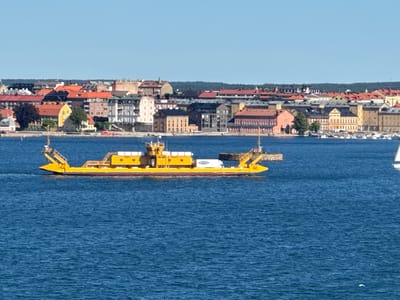 A yellow ferry boat, carrying a white truck, traverses the blue waters of a bay. In the background, the city of Karlskrona, Sweden, is visible. The city features a dense collection of buildings with red, orange, and yellow rooftops, interspersed with darker-colored structures and some green trees. The sky is a clear, bright blue, with no clouds present. The ferry appears to be moving from left to right in the frame. The water is choppy with small waves.