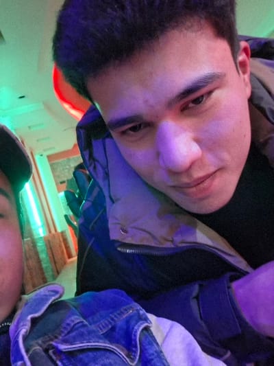 The image depicts a close-up, indoor scene, presumably within an entertainment venue in Takhtakuprik, Uzbekistan. The primary subject is a young male, positioned prominently in the right half of the frame. He has dark hair, dark eyes, and a fair complexion, with his gaze directed downwards and slightly to the viewer's right. His face is distinctly illuminated by a purple light source from the right. He is dressed in a dark puffer jacket over a dark undergarment.
A second individual is partially visible on the far left edge of the frame, showing only the side of their head, which is covered by a dark cap or similar headwear, and a portion of a blue denim jacket. This individual's visible area is illuminated by a green light. The background is out of focus, displaying blurred vertical elements and strong ambient lighting in shades of green, red, and orange, consistent with the interior of a casual gathering or entertainment establishment. The overall lighting scheme suggests a setting like a club, bar, or arcade.