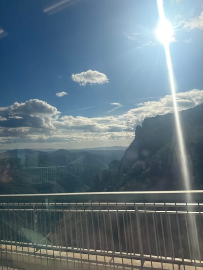 The image displays a panoramic view of the Vacarisses Viaduct (Viaducte de Vacarisses) in Vacarisses, Spain, under a bright, partly cloudy sky. The scene is observed from an elevated position, likely from a vehicle, indicated by a metal guardrail in the lower foreground. The sun, located in the upper right, emits a strong vertical flare across the image, casting intense light on the mountainous landscape.
In the midground, a deep valley is flanked by rugged, forested mountains, with discernible rock formations on the right. Spanning the valley on the left, the Vacarisses Viaduct is prominently visible, characterized by its multiple concrete arches or pillars supporting a broad roadway. The viaduct appears lighter in tone against the darker, shaded mountainsides. Scattered cumulus clouds and faint contrails are present in the blue sky. Distant mountain ranges form the hazy horizon. No individuals, specific actions, or interactions are observable.