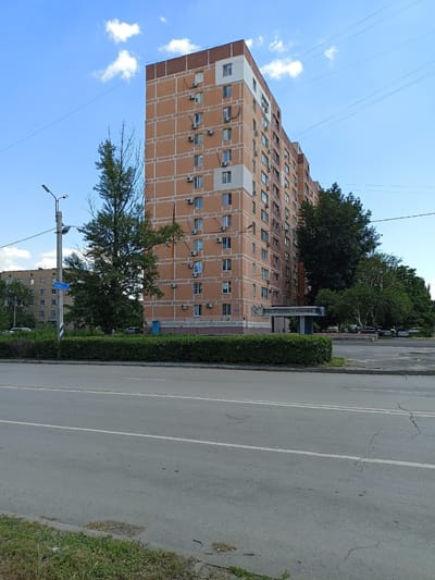The image depicts a wide asphalt road in front of a multi-story residential building in Volgodonsk, Russia. The building is orange with white trim and has numerous windows and air conditioning units. A light pole with a street sign that reads "ул. Ленина" (Lenin Street) is visible to the left of the building, along with a trimmed green hedge. A large tree stands between the street sign and the building. In the background, more buildings and trees are visible, as well as a parking lot with several cars. The sky is blue with scattered white clouds. The overall scene is that of a typical urban environment on a sunny day.