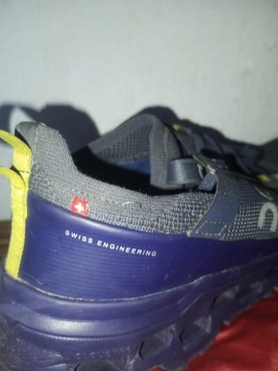 A navy blue running shoe with a distinctive design, featuring the words "SWISS ENGINEERING" printed in white on the side of the sole, and a Swiss flag above the text. The upper portion of the shoe is a textured gray material, and a yellow pull-tab is visible at the back. The sole of the shoe has a distinct pattern. The background is a blurry mix of white and red, adding to the close-up perspective of the shoe.