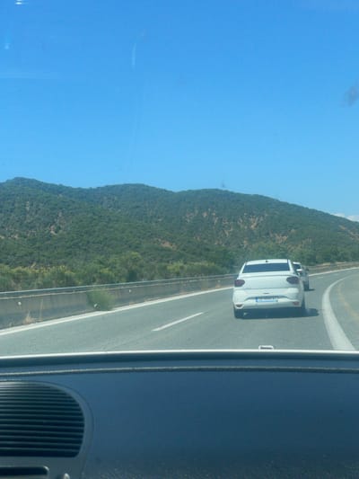 The image displays a daytime highway scene from the interior perspective of a moving vehicle. The bottom portion of the frame shows a dark-colored dashboard, featuring a black, horizontally-ribbed grille on the left side, and the lower edge of the windshield.
Ahead, a multi-lane asphalt highway extends into the distance, exhibiting clear white lane markings and a gradual curve to the right. A concrete barrier with a metal guardrail runs along the left side of the road, with sparse dry vegetation growing beside it.
Two vehicles are visible on the highway in front of the camera. The primary subject is a white Dacia Logan sedan, positioned in the right lane. Its rear is distinctly visible, displaying a blue Greek license plate with the alphanumeric identifier "CB 9840 AO". Behind this vehicle, in the left lane, another light-colored sedan is partially observed further along the road.
The background features a expansive range of hills or mountains, densely covered with green trees and vegetation, under a clear, bright blue sky. No human figures are discernible. The "CB" prefix on the Greek license plate corresponds to the Kavala regional unit, consistent with the specified location of Δημοτική Ενότητα Προμαχώνος, Greece, given its proximity to the Serres regional unit.